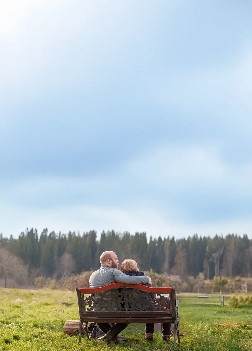 Two people sitting together on a bench overlooking open land and trees at Rooted Northwest.