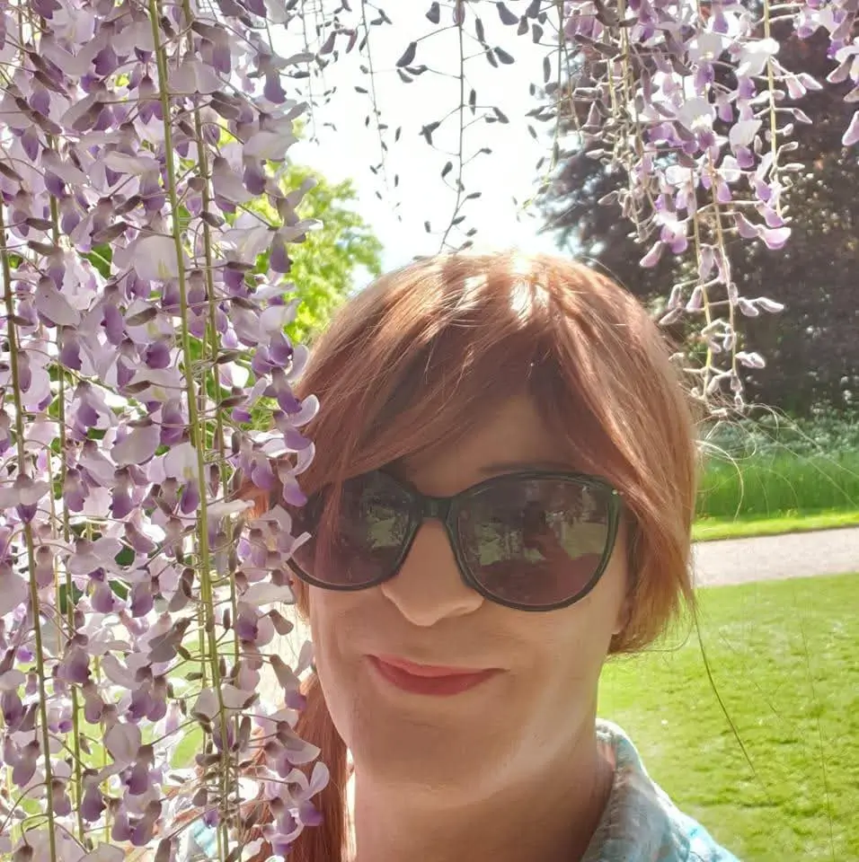 A person with sunglasses smiles in front of cascading purple flowers, surrounded by greenery and sunlight.