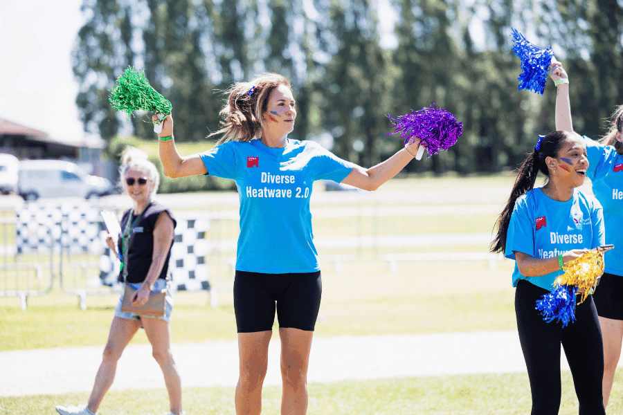 Outdoor team-building event with participants wearing matching blue shirts, taking part in inflatable obstacle courses and games, smiling and cheering, with the words 'ENTER A TEAM' in large bold white and green text across the image.