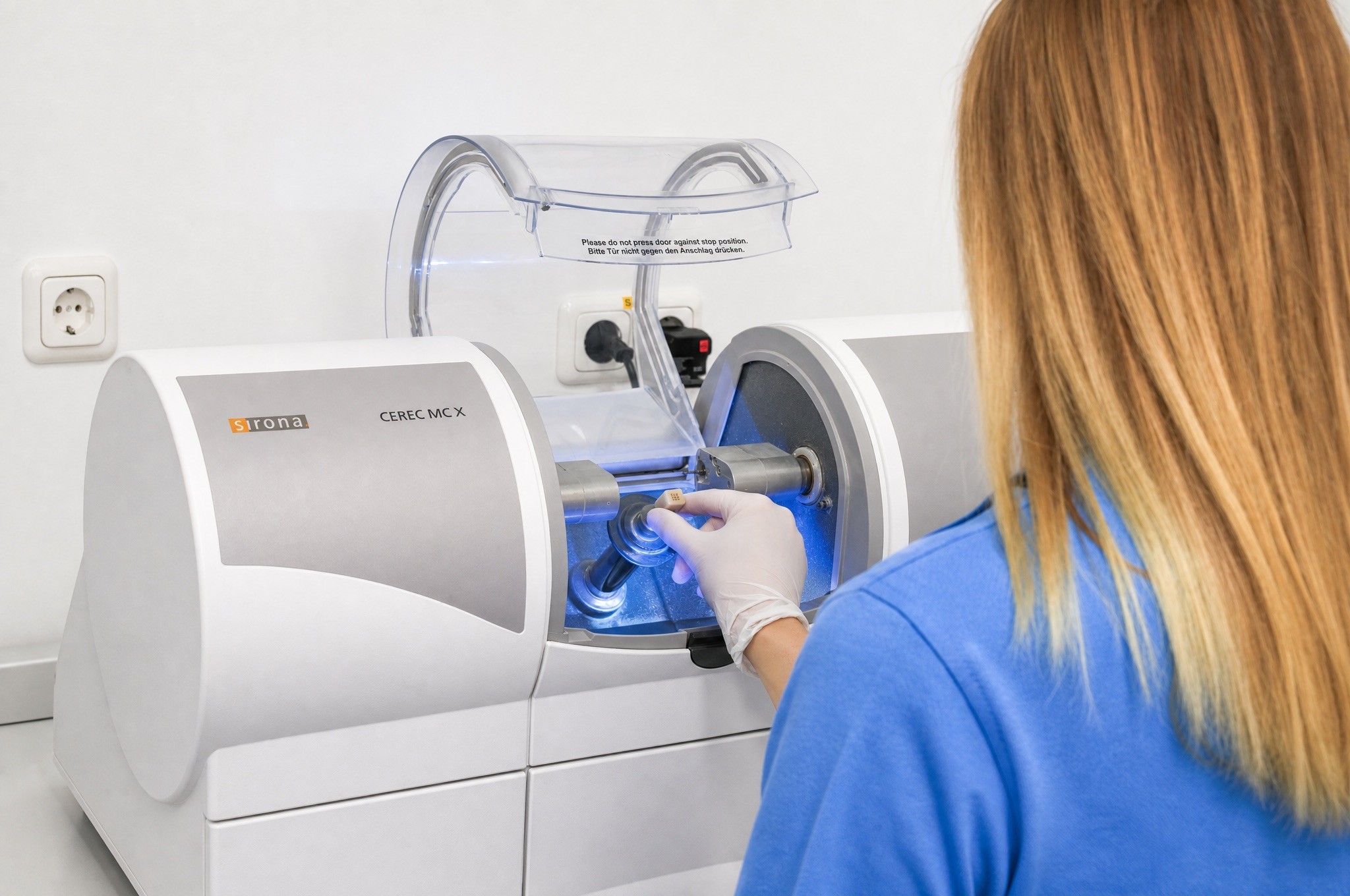 A dental assistant with a blonde ponytail and a blue polo shirt operating a precise, modern white dental milling and laboratory machine.