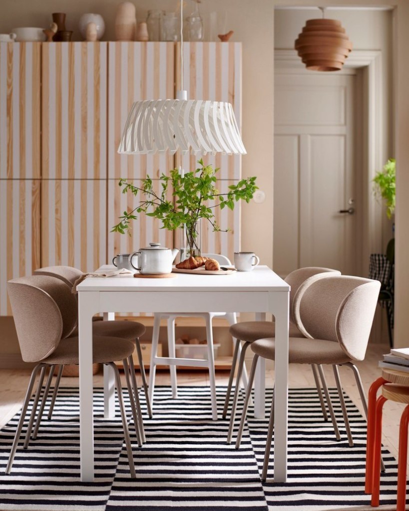 A modern dining room featuring a white table surrounded by beige chairs, with a decorative lamp hanging above. The table is set with teacups, a teapot, and croissants, complemented by a vase of greenery. The backdrop is a stylish cabinet with vertical striped design and a striped rug on the floor.