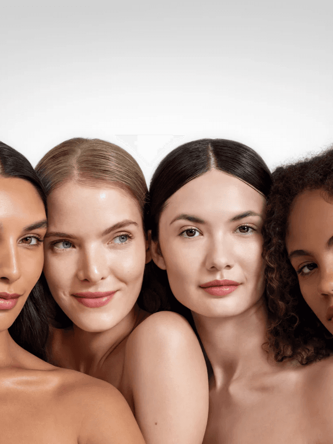 Four women of diverse backgrounds with natural makeup, showcasing different skin tones and hair textures, smiling against a neutral gray background.