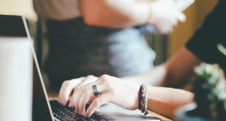 Imagem focada nas mãos de uma pessoa digitando em um laptop. A pessoa usa uma aliança escura e algumas pulseiras coloridas no pulso, o que adiciona um toque casual ao ambiente de trabalho. Ao fundo, uma outra pessoa está desfocada, criando uma sensação de colaboração em um espaço compartilhado, como um escritório ou coworking. A imagem transmite uma atmosfera descontraída e moderna, ideal para representar ambientes de trabalho flexíveis ou de tecnologia.