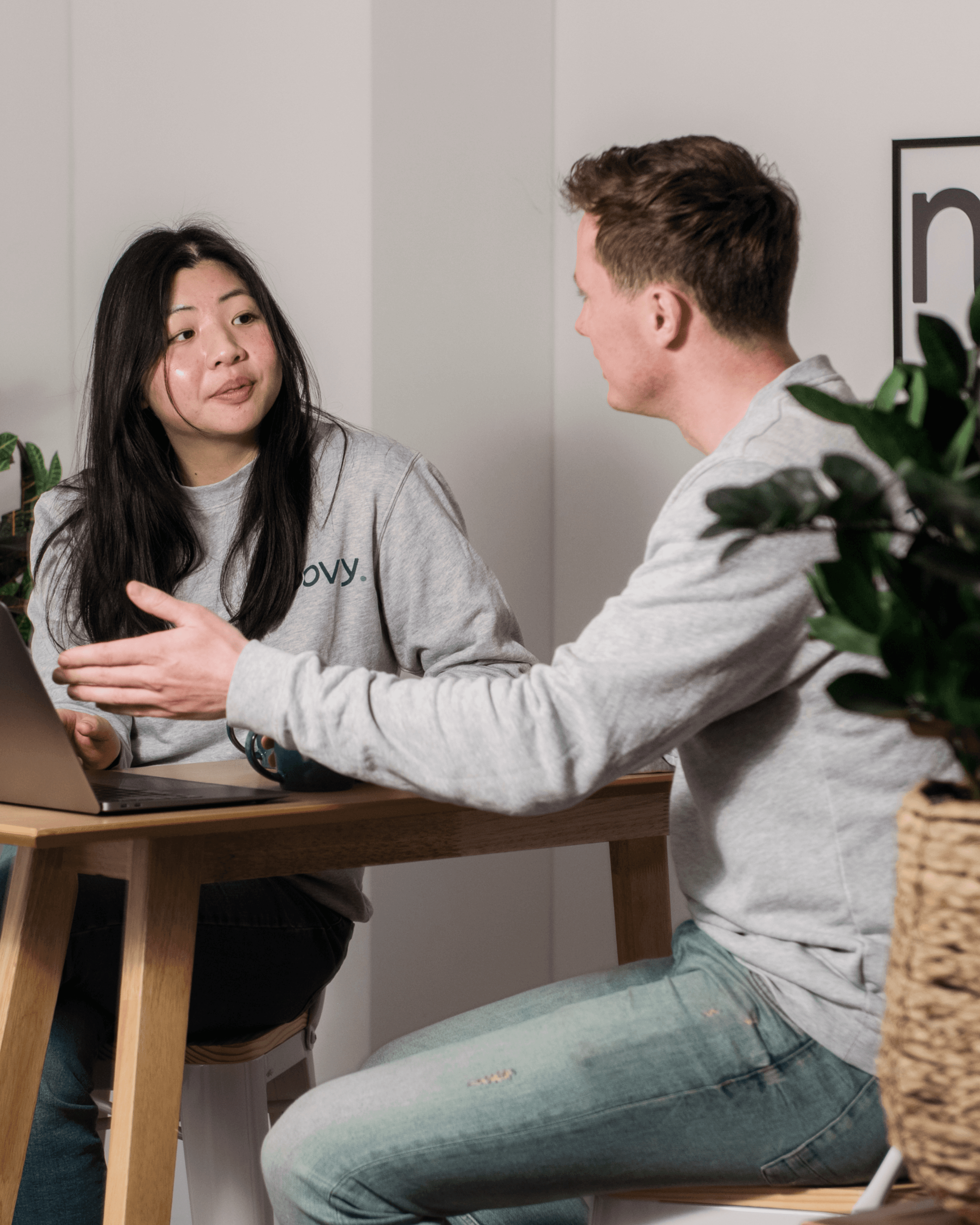 A young woman and man engaged in a conversation at a table, surrounded by indoor plants.