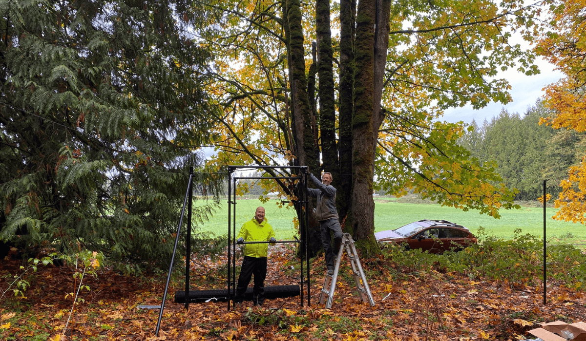 Two volunteers secure fencing near a large tree as fall leaves cover the ground at Rooted Northwest.