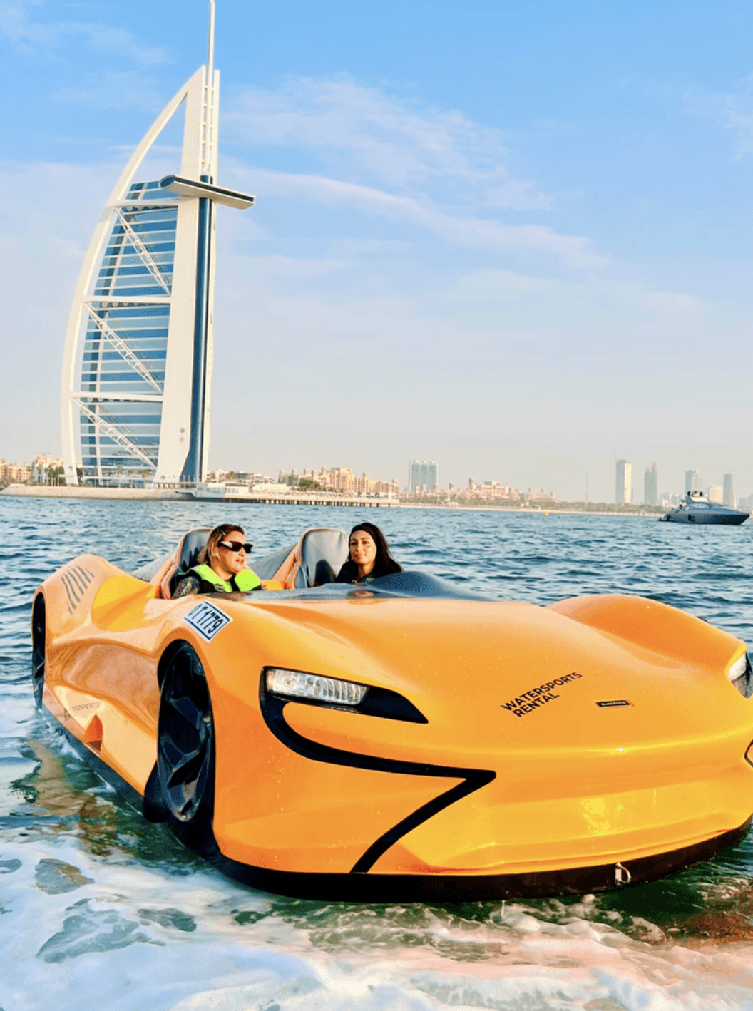 Two women riding a jet car with Dubai's iconic sail-shaped building in the background.