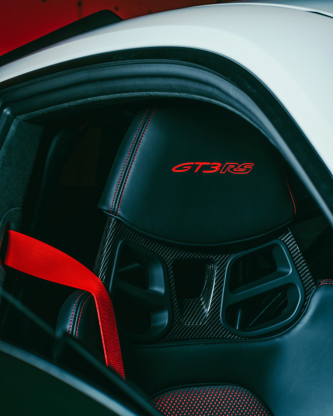 Detail of a white Porsche 911 GT3 RS (992) interior through the side window, showing the carbon-fibre bucket seat with red "GT3 RS" embroidered headrest and red six-point harness.