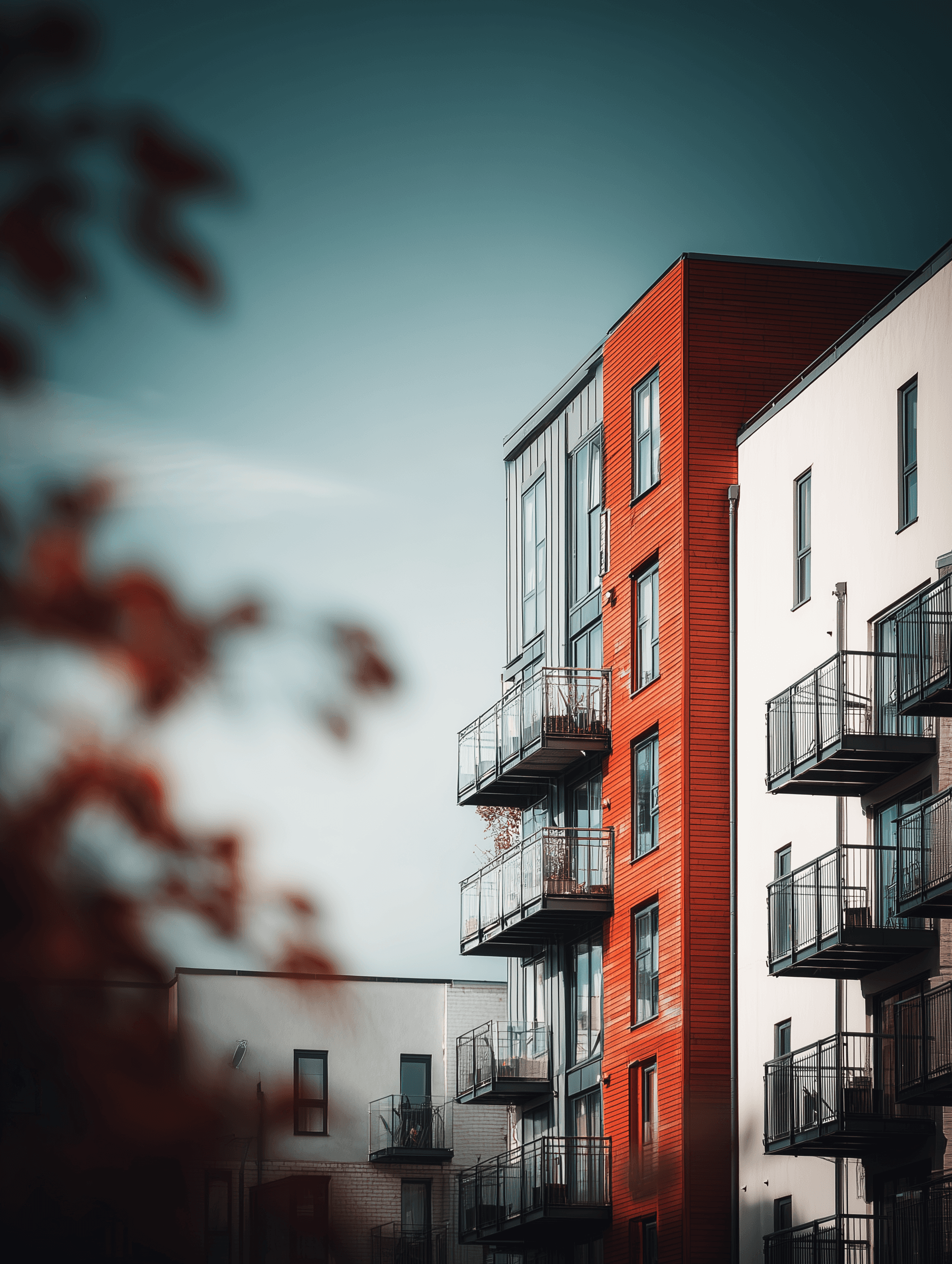 Photograph of modern UK build-to-rent apartment buildings featuring a striking red facade section with multiple glass balconies. Surrounding buildings have white and grey exteriors. The image is softly framed with blurred foliage in the foreground and set against a clear, teal-toned sky