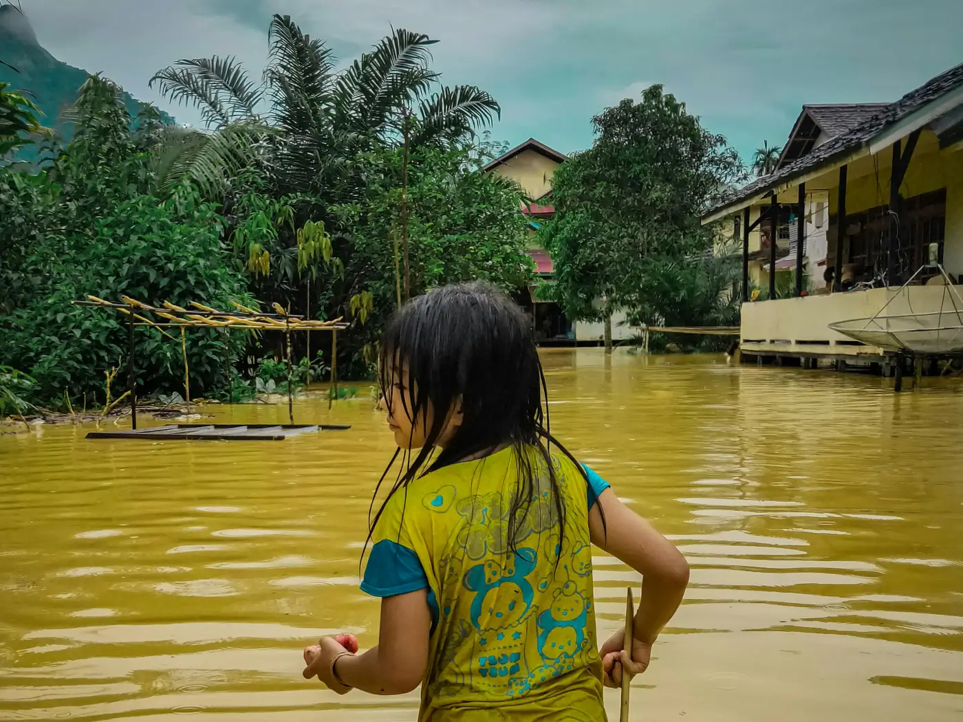 A child wades through muddy floodwaters, surrounded by damaged trees and houses.
