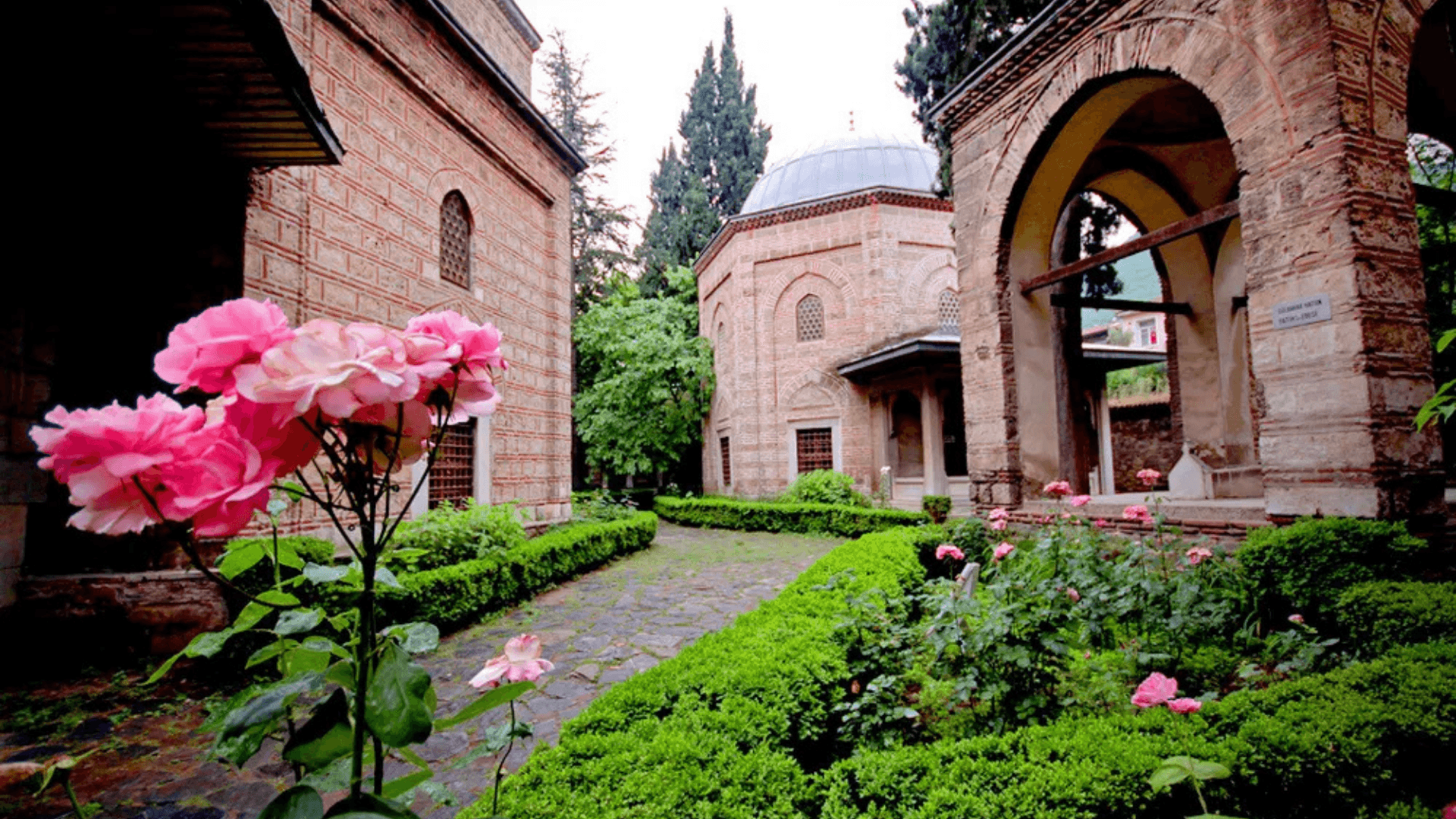Historic Ottoman courtyard garden with blooming roses, trimmed hedges, and traditional brick buildings in Bursa, Türkiye