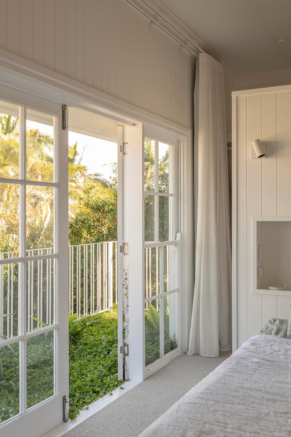 Bedroom opening to a balcony through glazed doors, framed by soft curtains and garden views beyond.