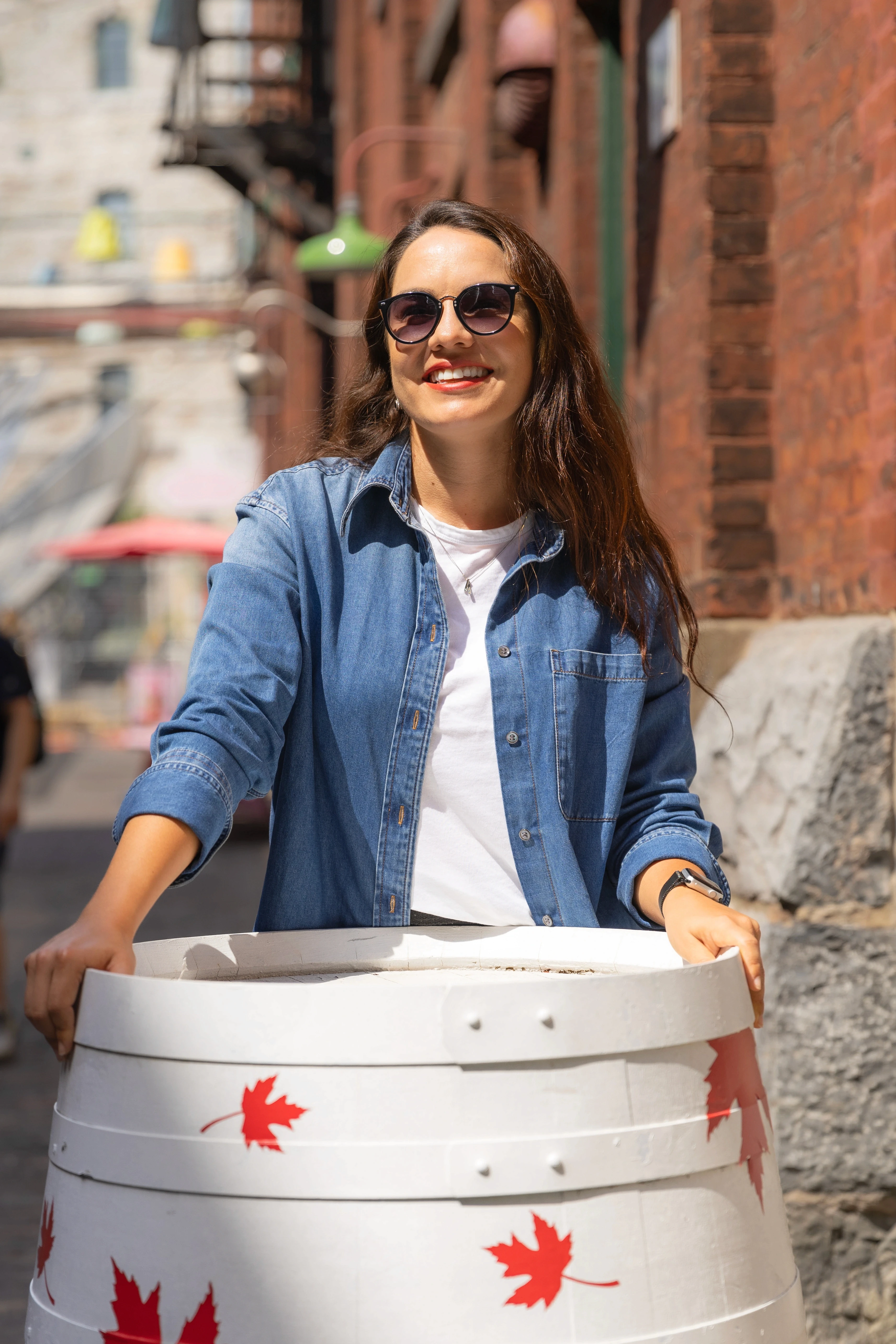 Mulher sorrindo em rua histórica de Toronto, encostada em um barril branco com folhas de bordo pintadas.