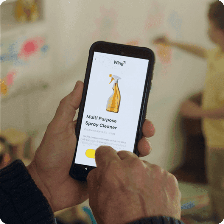 A person is holding a smartphone displaying an app featuring a multipurpose spray cleaner, with a blurred background of a room and a child.