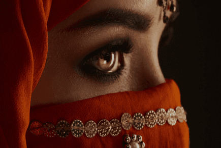 Gros plan sur le regard d’une femme indienne portant un voile orange sombre orné de pièces dorées, ambiance mystérieuse et élégante