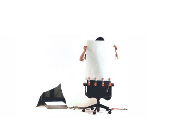 A person holds a large white sheet of paper in front of their face, obscuring it. They are standing behind a black rolling chair with orange straps attached to its back. A black triangular object with a white surface sits on the floor to the left of the chair. A black cord extends from the chair toward the object. The background is plain white.