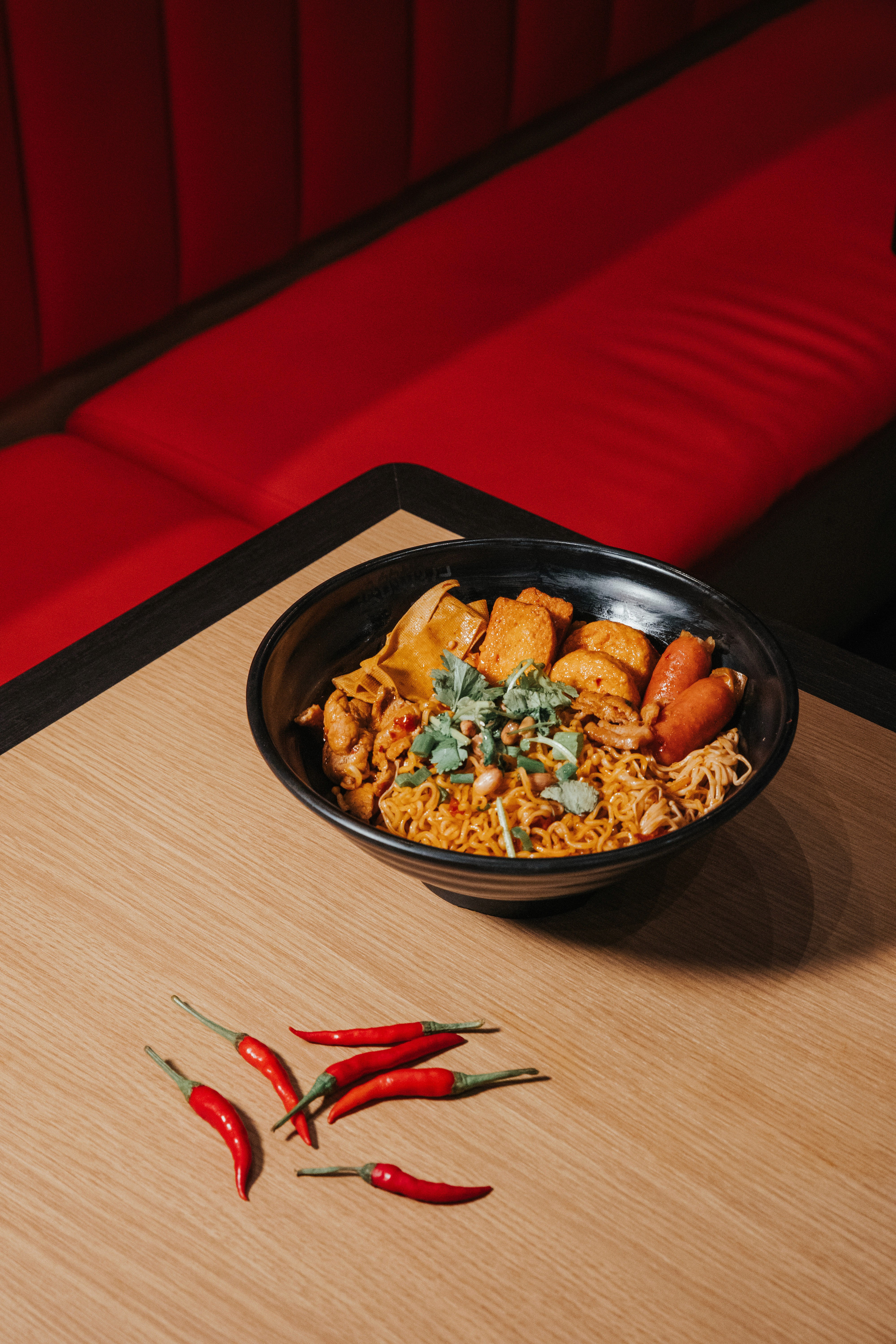 A bowl of colorful noodles garnished with herbs, placed on a wooden surface, with red chilies nearby.