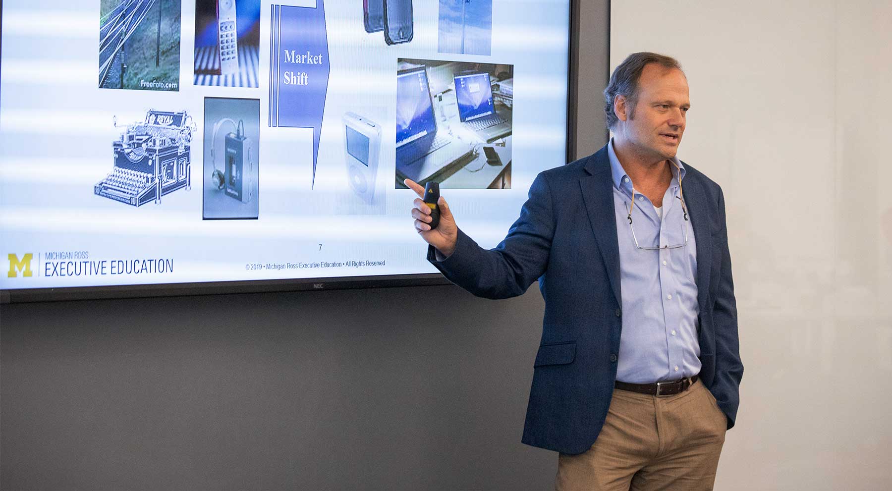 A presenter stands in front of a screen displaying visuals, gesturing while explaining a topic.