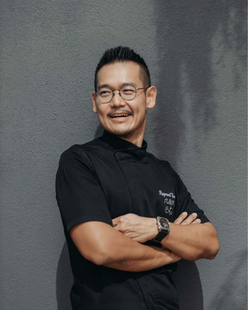 Chef Raymond Tham poses with arms crossed, wearing a black culinary uniform and round glasses, smiling against a grey wall.