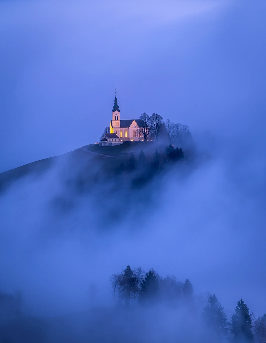 A church in Slovenian countryside schrouded in fog during blue hour.