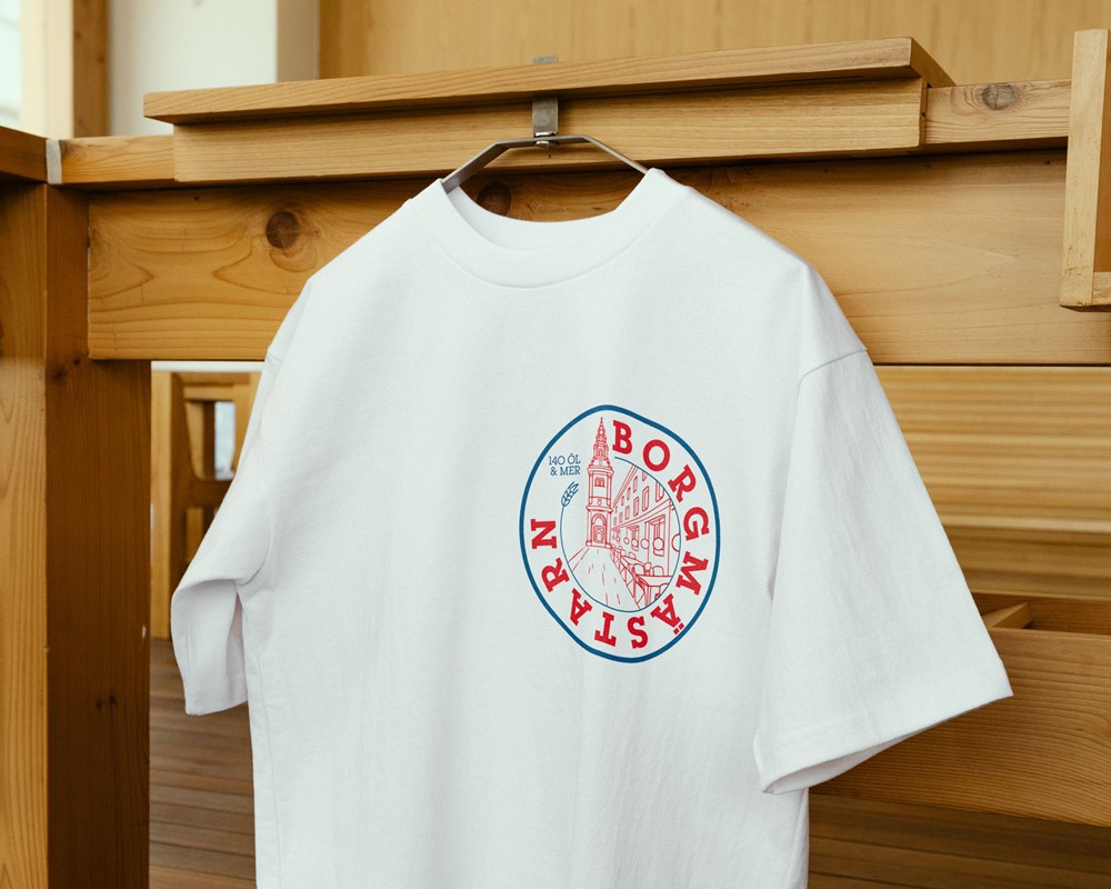 A white t-shirt with the Borgmästarn red circular logo hanging on a wooden hanger against a warm neutral background, part of the pub's branded merchandise range.