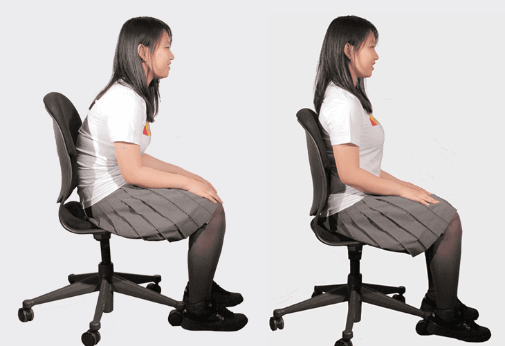 A woman with dark hair sits in a black office chair with rolling casters. She wears a white short-sleeved shirt and a gray pleated skirt with black tights or stockings. Her legs are crossed at the ankles, and she looks to her left. The background is plain white.