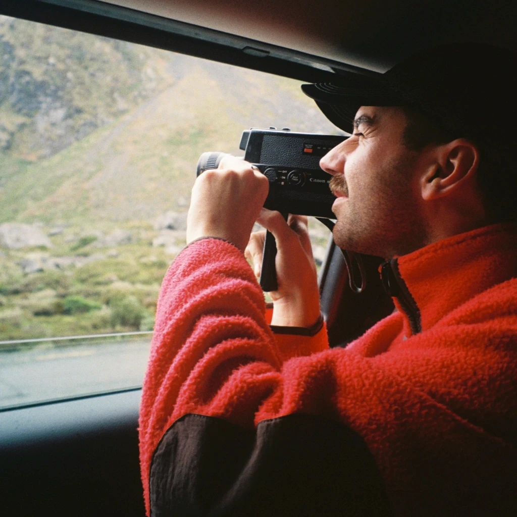 Lachy Henry - wedding videographer - in a red jacket uses a camcorder to film the scenic, lush green mountain landscape outside a car window.