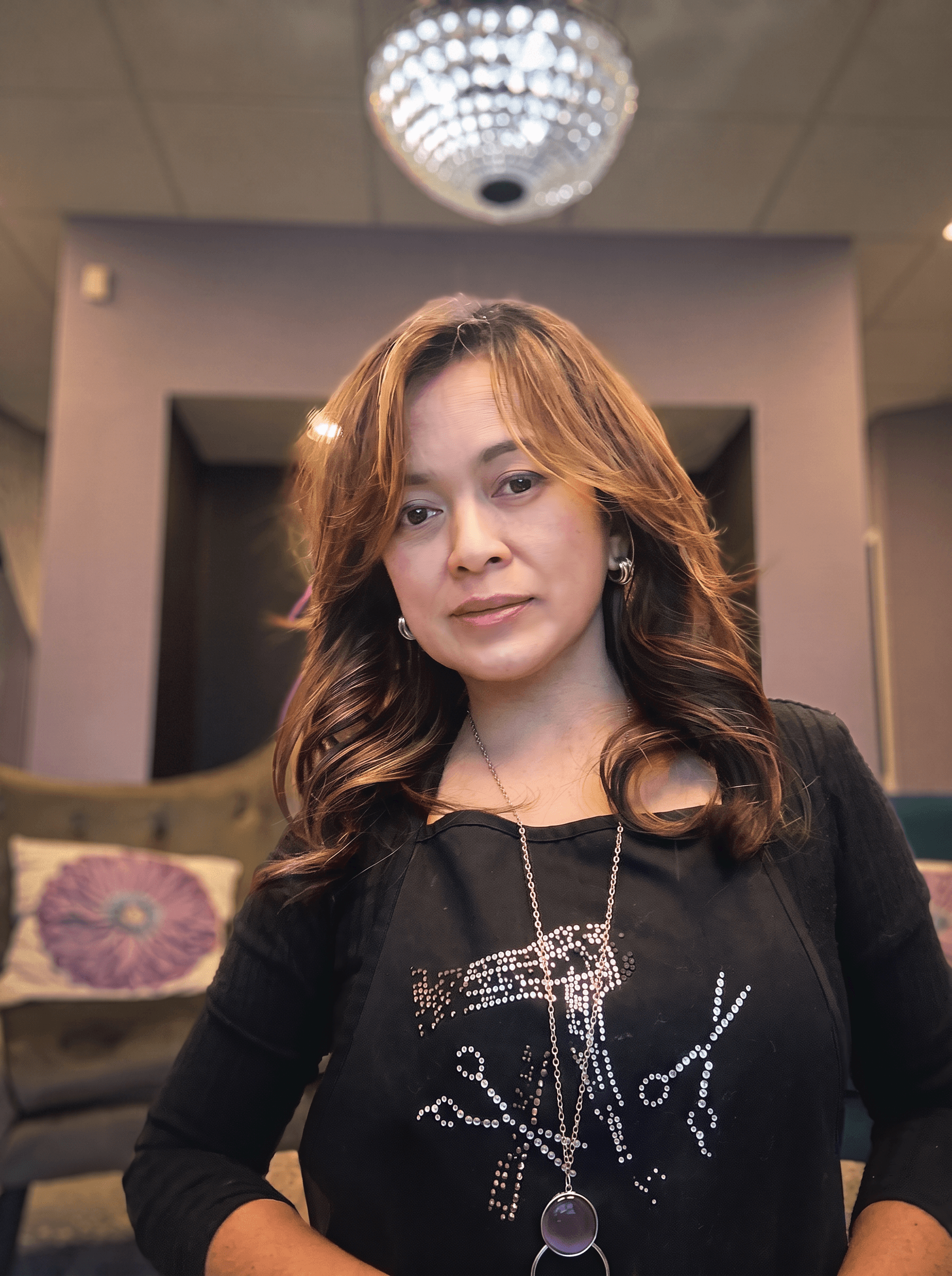 Professional portrait of a smiling woman with dimensional caramel-brown hair, wearing a salon apron with a scissor graphic and a crystal chandelier in the blurred background.