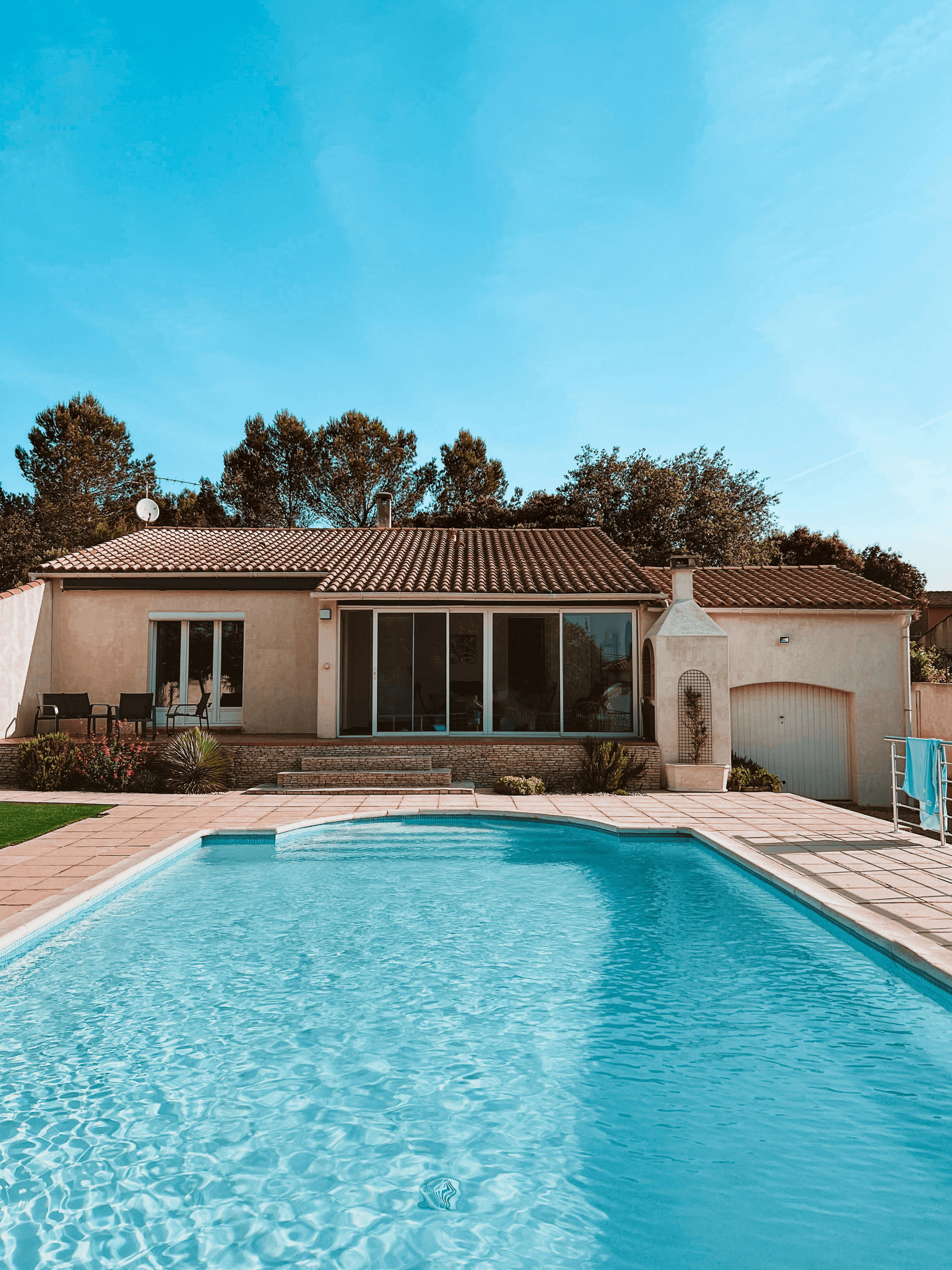 Modern luxury house with beige stucco exterior, terracotta roof tiles, and a rectangular turquoise pool in the front yard, set against a bright blue sky.

