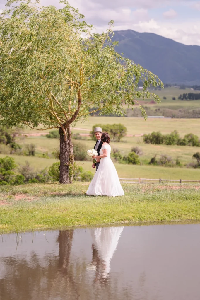 A scenic shot of the bride and groom walking near a reflection pond with a lone willow tree and sweeping valley views at a Chaparral Ranch Wedding.