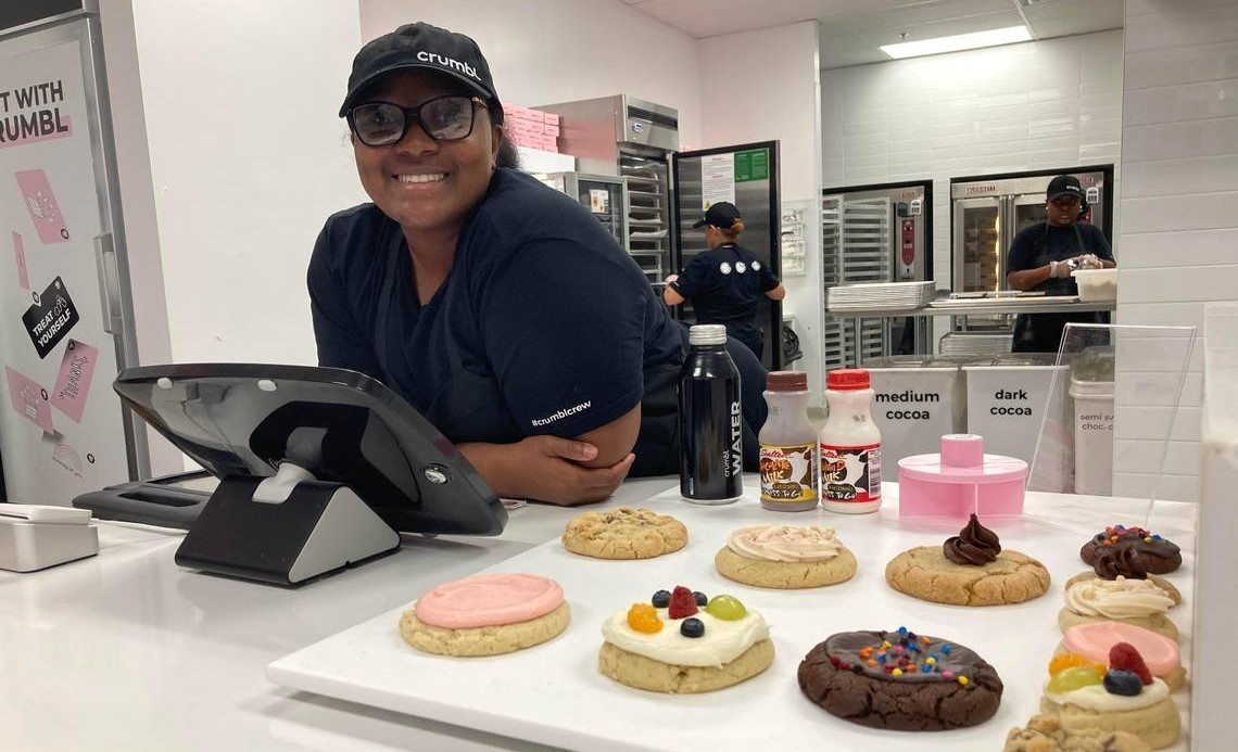 Une employée Crumbl Cookies pose derrière un plateau de cookies décorés, dans un décor rose et blanc