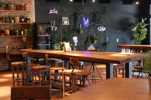 Großzügiger Gemeinschaftsbereich im kAlps mit langen Holztischen, Hockern, Pflanzen und dekorativen Neonlichtern.