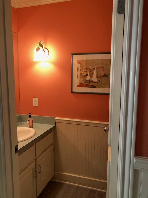 A small bathroom features vibrant orange walls, a single wall-mounted light, a framed sailboat artwork, a green ceramic sink with a soap dispenser on a light-colored countertop, and cream-colored cabinetry amidst wood-paneled wainscoting.