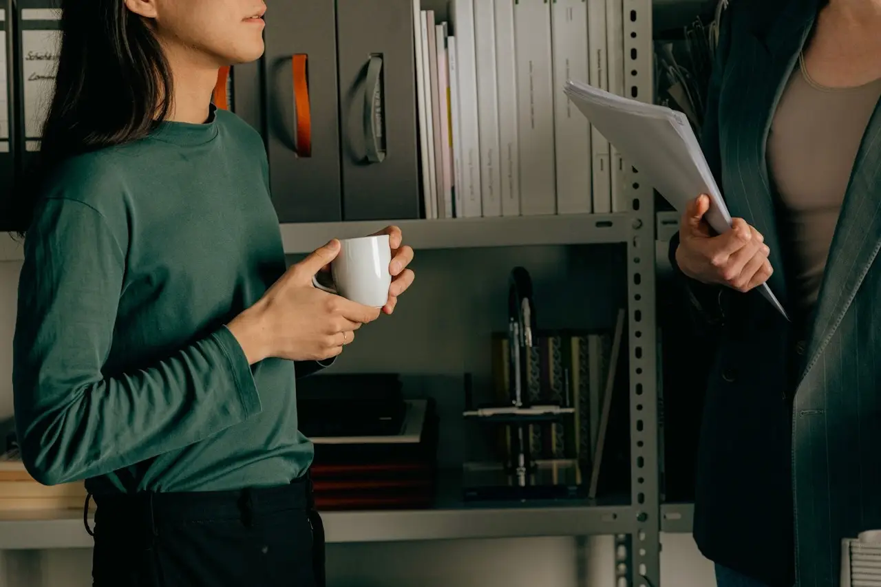 close-up of a person’s hand holding a white mug in an office setting with blurred bookshelves in the background.
