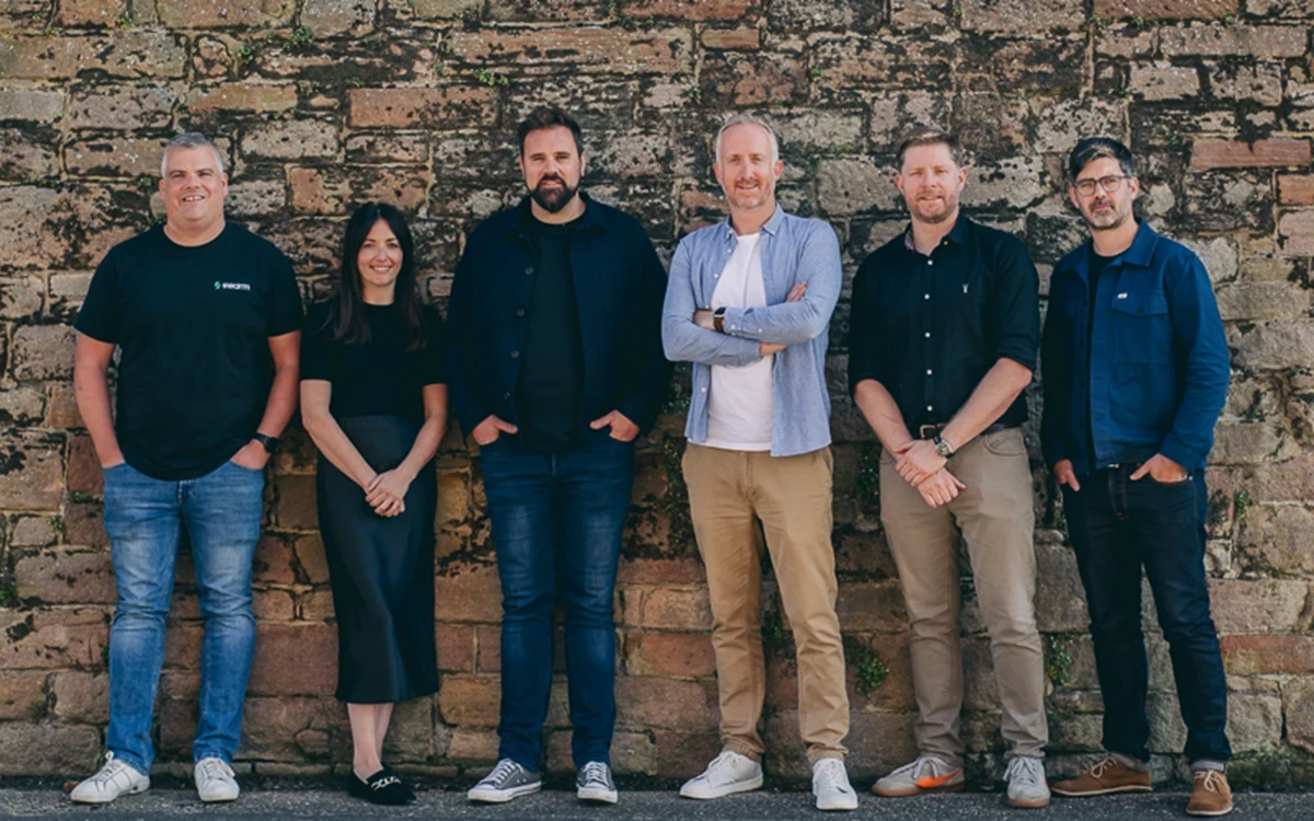 A group of 6 people standing in a line against an old brick wall looking at the camera