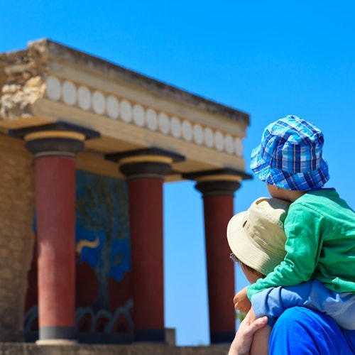 An adult carrying a child on their shoulders looks at ancient ruins with red columns and detailed frescoes under a clear blue sky.