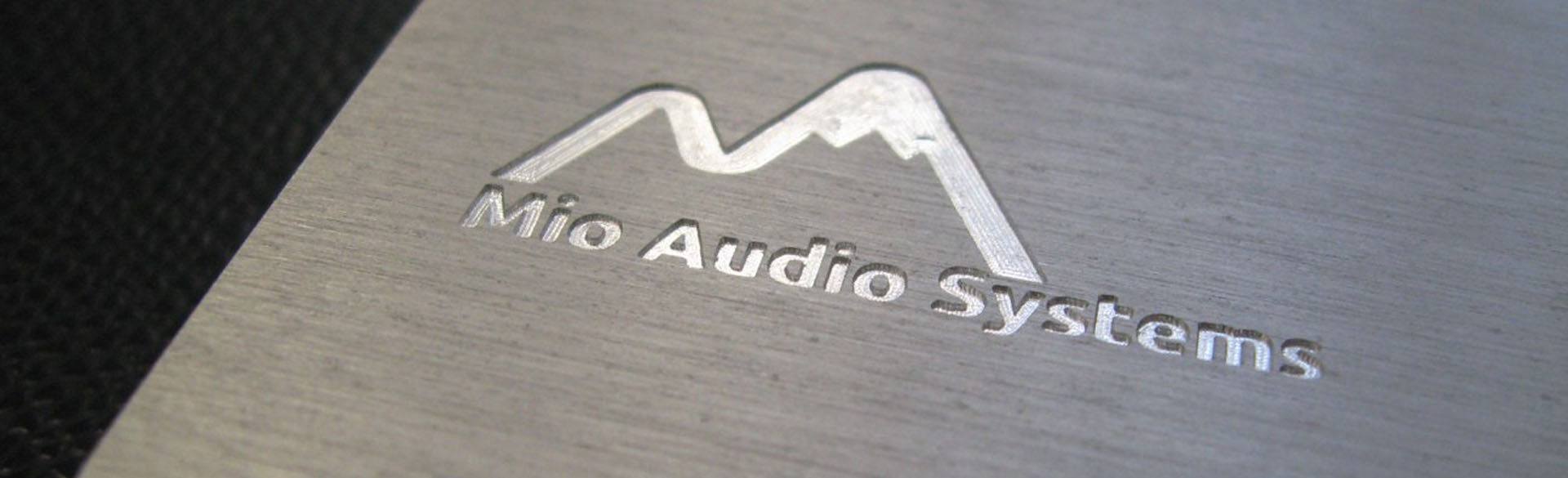 Logo métallique argenté « Mio Audio Systems » avec une forme de montagne stylisée, gravé sur une surface métallique brossée à côté d'un matériau noir texturé.