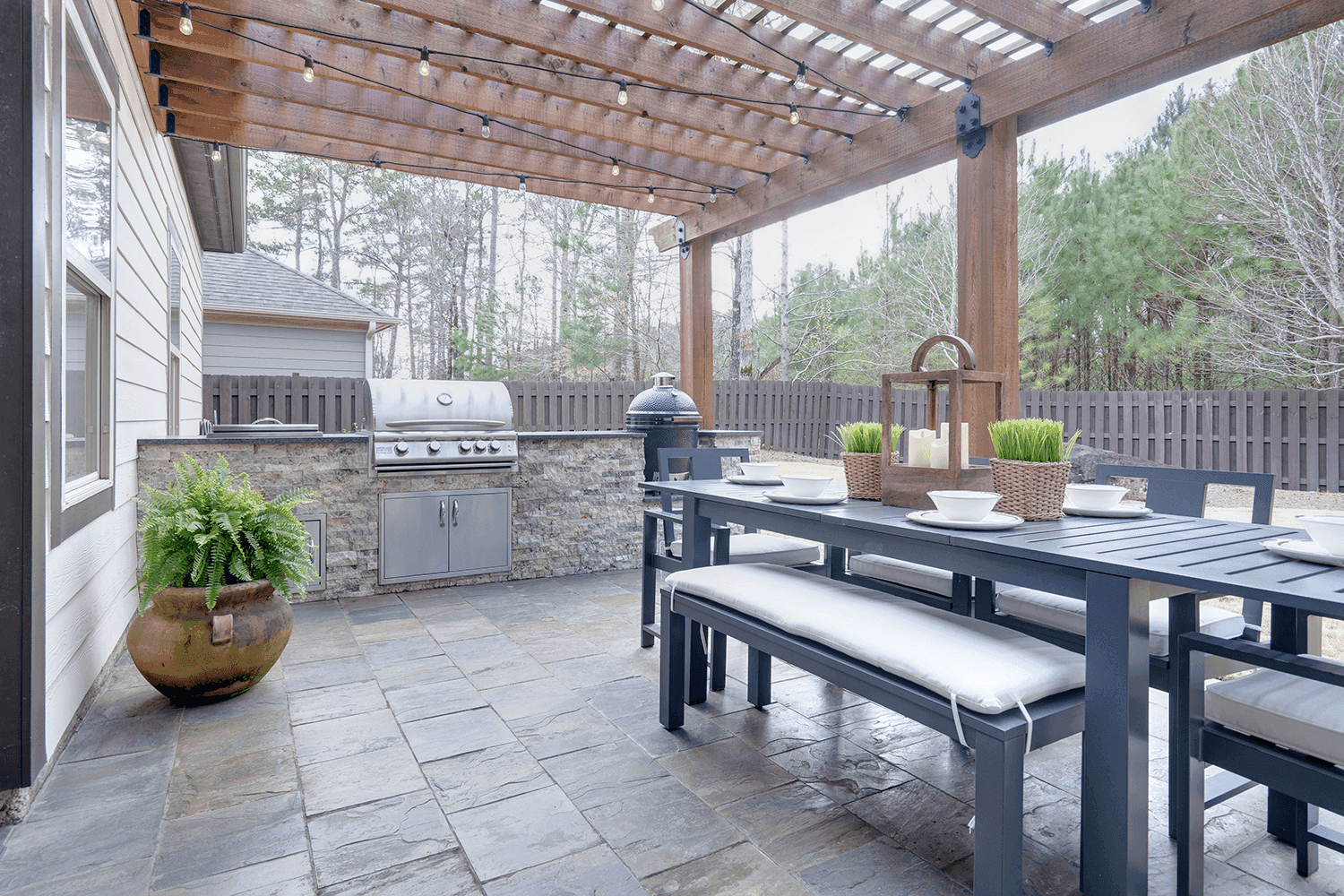 A sleek outdoor kitchen with a stainless steel grill and stone-clad base, positioned under a wooden pergola strung with warm string lights. Nearby, a black dining set with white cushions is arranged, featuring decorative plants and table settings, creating a modern, inviting patio space.