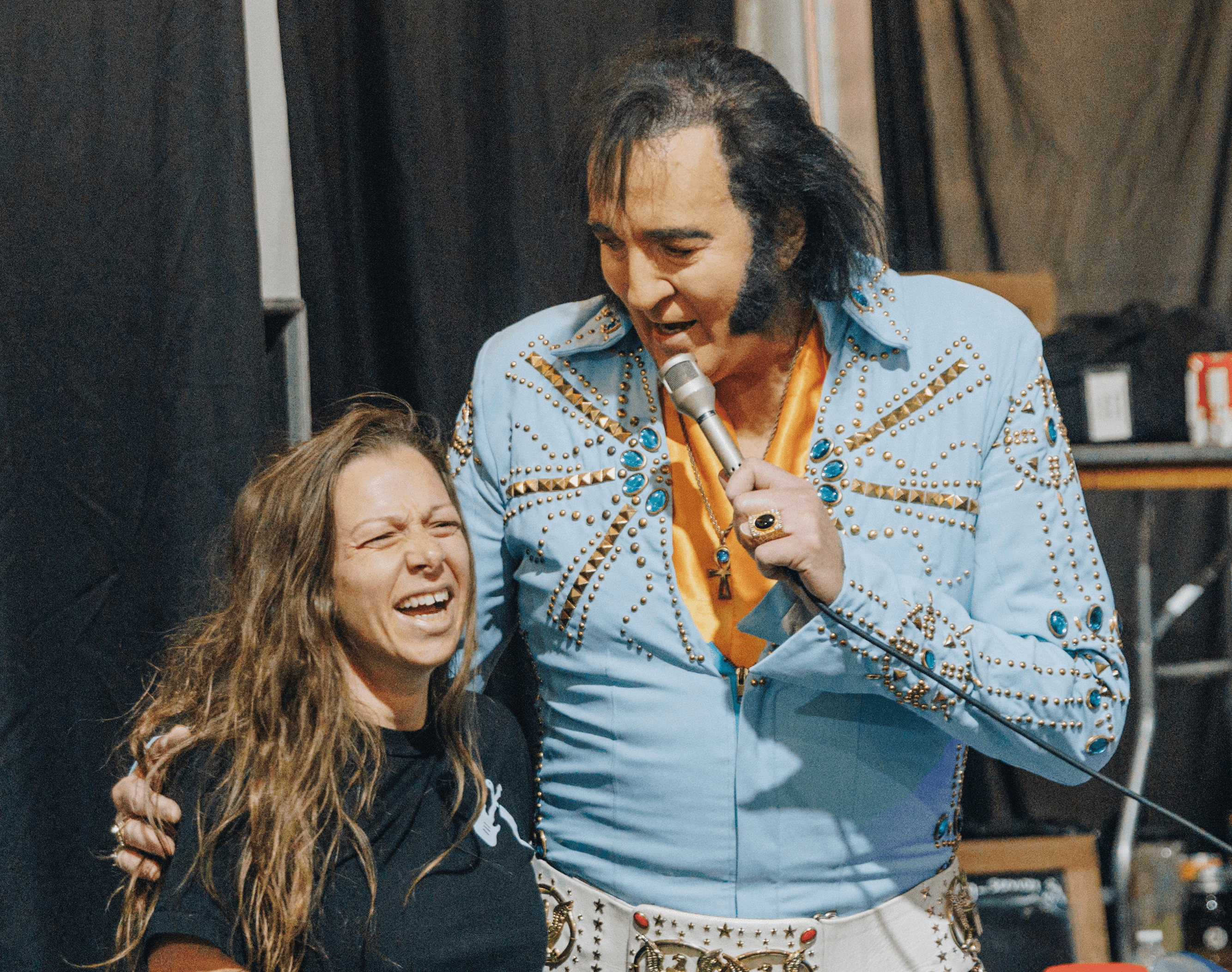 A smiling woman and a man in a blue outfit stand together, engaging in conversation at an event.