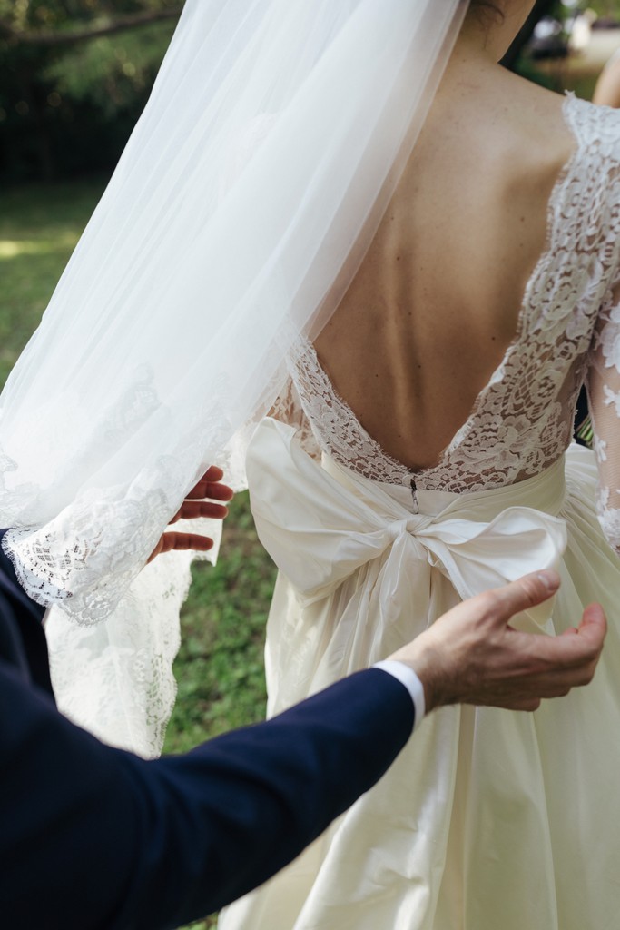 Sposo accarezza il fiocco di seta sulla schiena dell'abito da sposa, dettaglio di pizzo e tulle nel giardino della villa