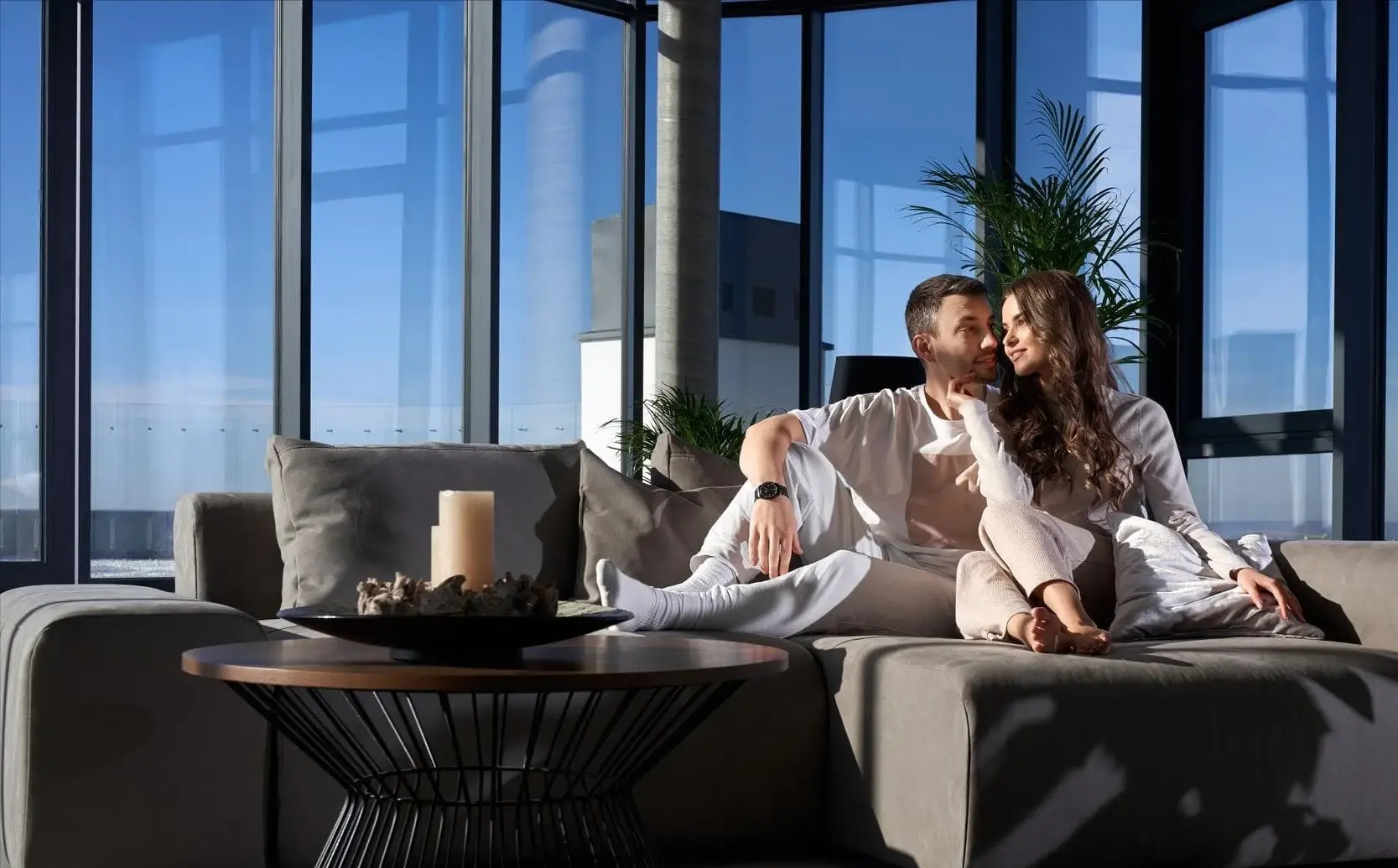 A couple relaxes together on a modern gray sofa in a luxury apartment with floor-to-ceiling windows and city views. The image conveys comfort, success, and the lifestyle made possible through Jumbo Loans from Chris Lewis Home Loans.