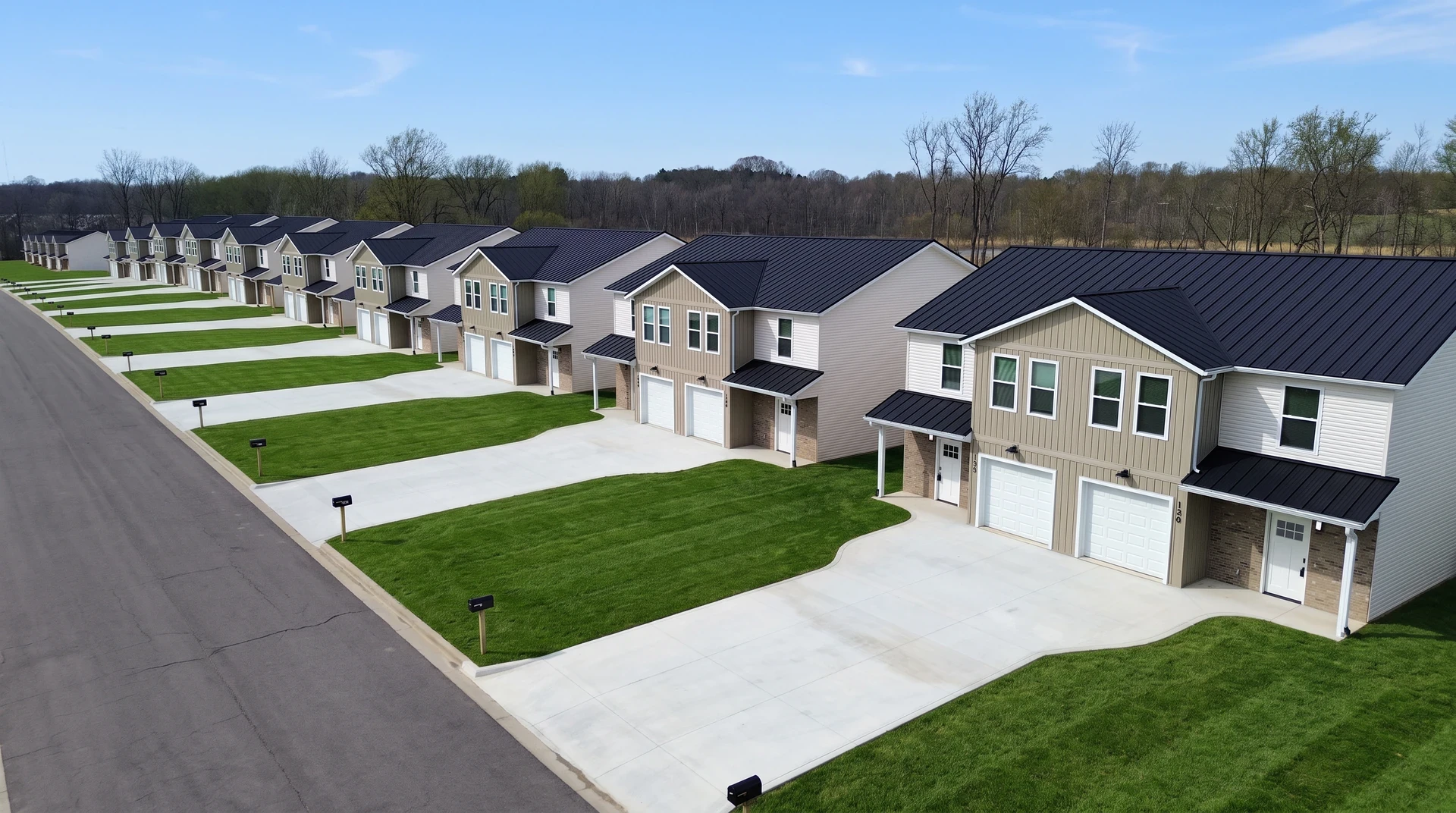 Aerial view of a long row of tan and white two-story duplex homes with dark standing-seam metal roofs, white garage doors and individual concrete driveways, backing onto a bare-tree treeline