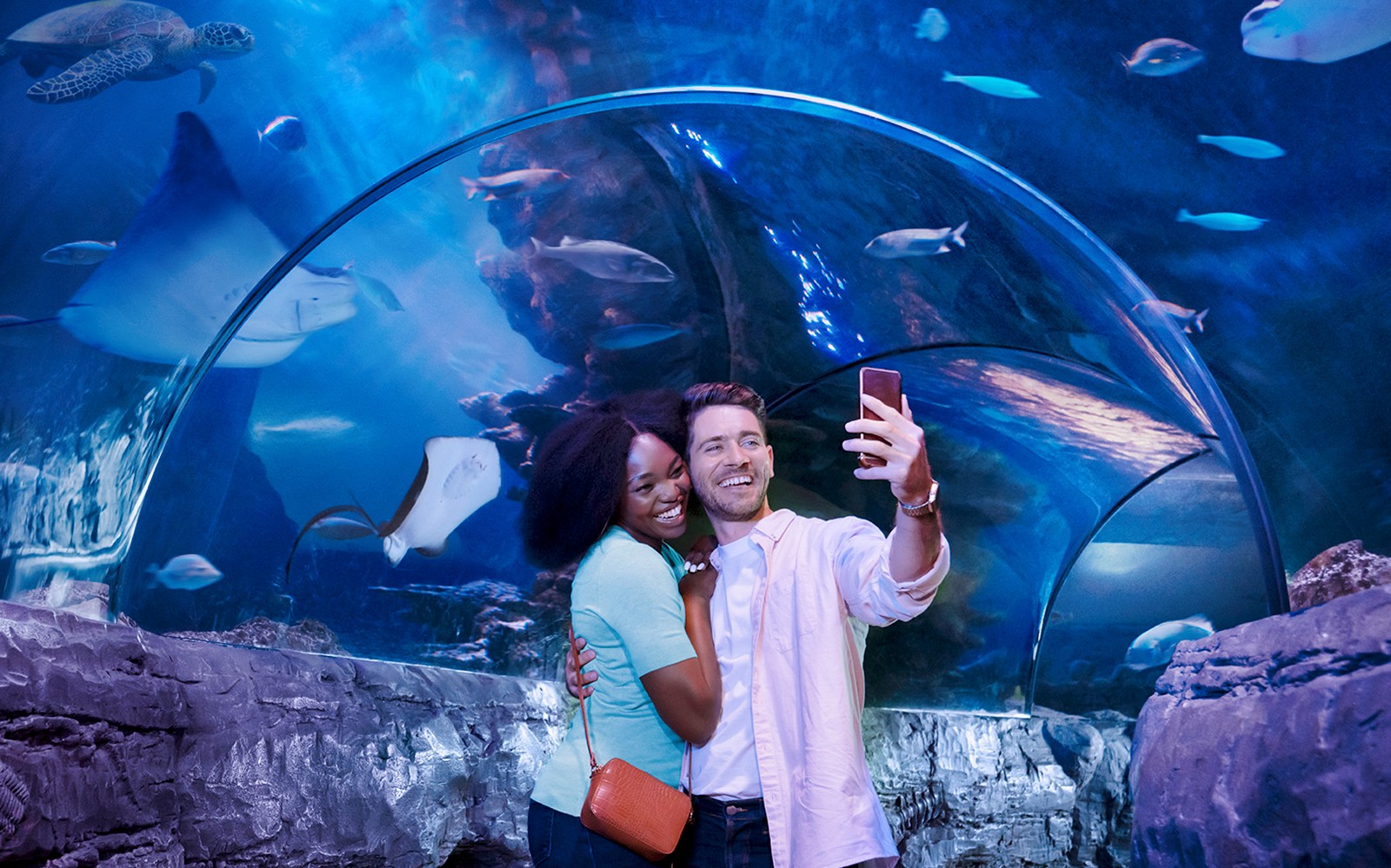 Tourists taking a selfie in an aquarium tunnel with fish and rays swimming above.