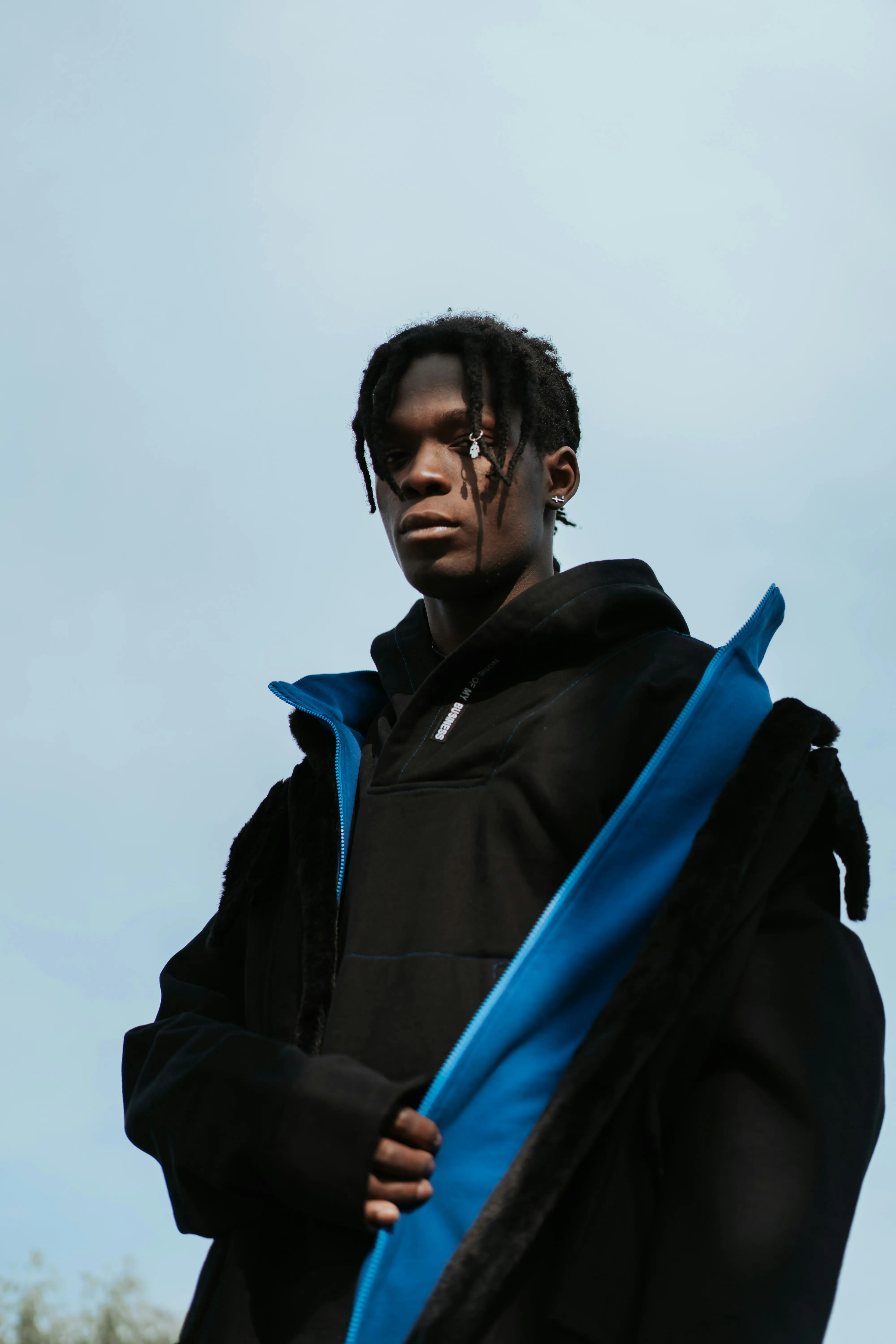 A low-angle portrait of a young man with braided hair, wearing a black hoodie with a vibrant blue jacket, set against a pale blue sky.