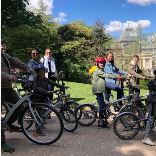 Mavi gökyüzü altında arka planda bir bina ile yeşilliklerle dolu bir parkta, aralarında çocukların da bulunduğu bir grup insan bisiklet sürüyor.