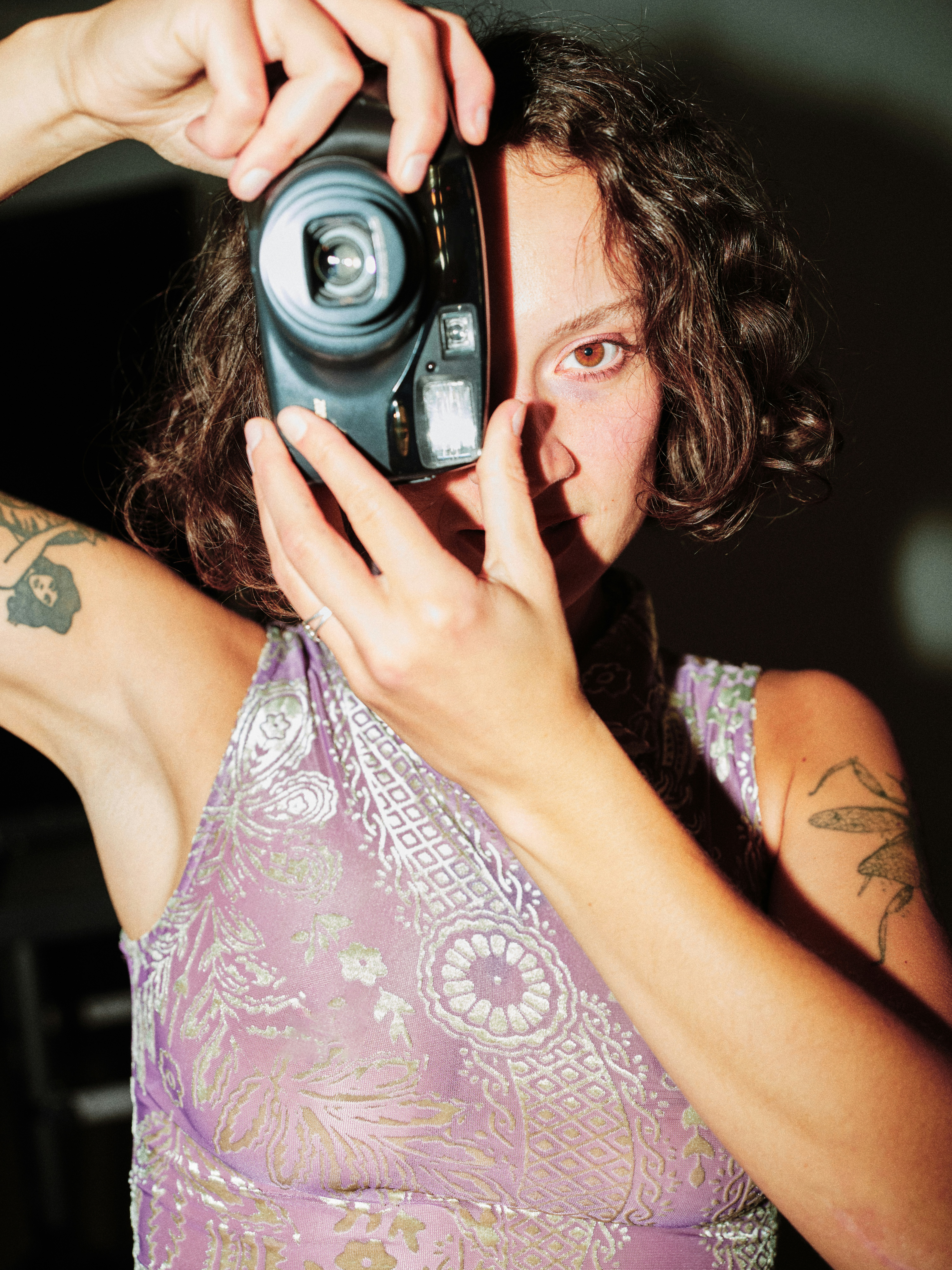 Close-up editorial portrait of a tattooed model holding a film camera up to her face, wearing a purple metallic patterned top, dramatic dark background