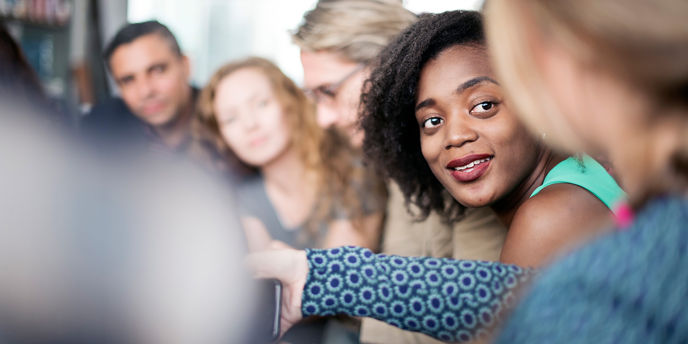 A woman wearing a turquoise top gestures while speaking during what appears to be a workplace meeting or training session. She is in sharp focus in the foreground, with several diverse colleagues visible but blurred in the background, suggesting an engaged group discussion or collaborative work environment.