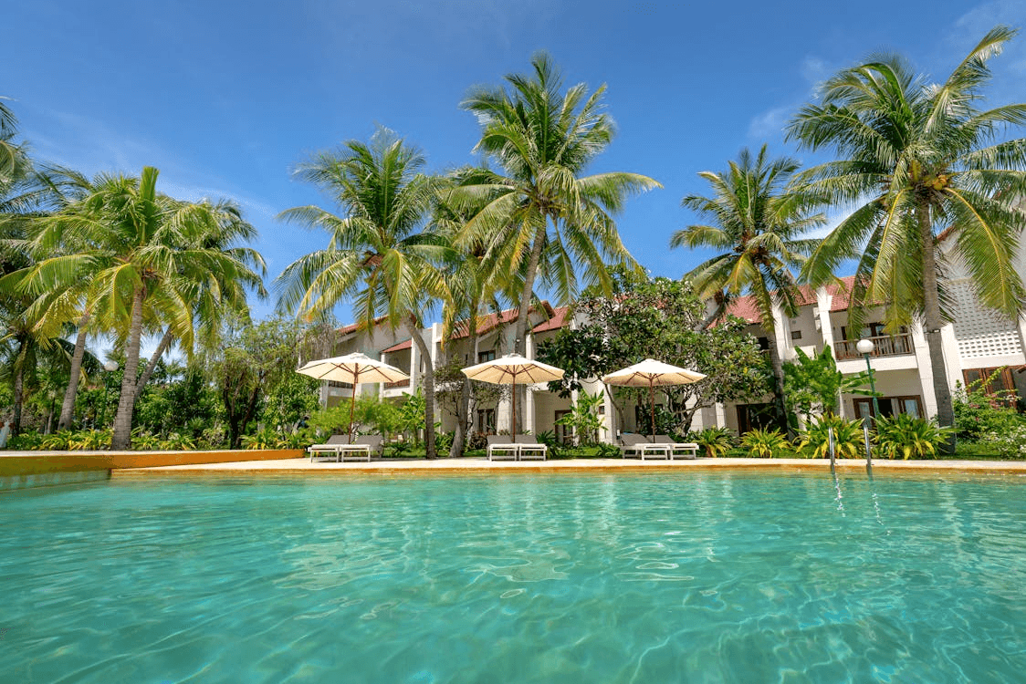 Red-roofed tropical resort building with tall white columns, palm trees, and large rectangular pool with lounge chairs.

