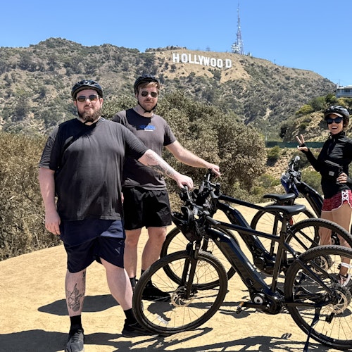 Three people pose with bicycles in front of the Hollywood sign on a sunny day.