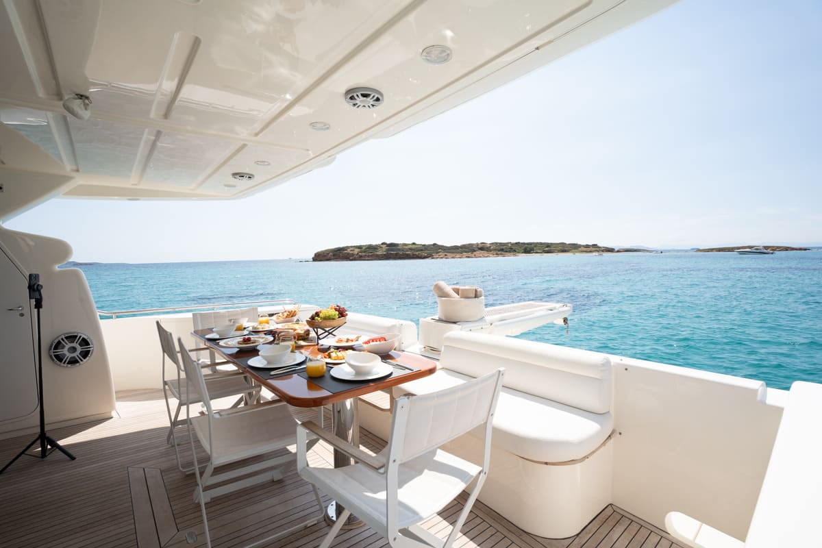 Ferretti 63 yacht's outdoor dining area with breakfast spread on teak deck, overlooking turquoise waters and small islands in the Cyclades.