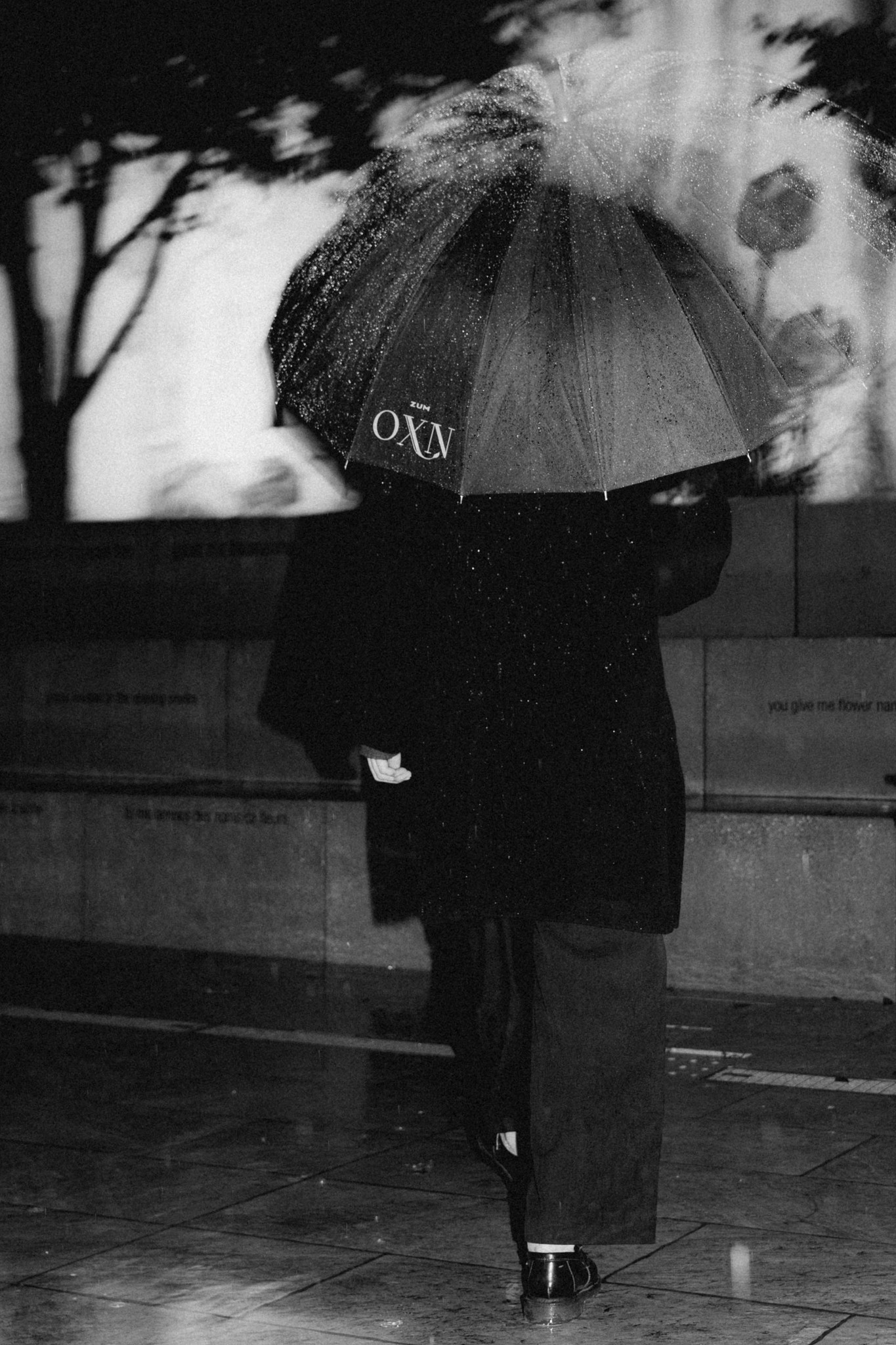 Schwarz-Weiß-Fotografie einer Person, die im Regen mit einem 'zum OXN'-Regenschirm geht, mit dem Schriftzug 'RAINDROPS'